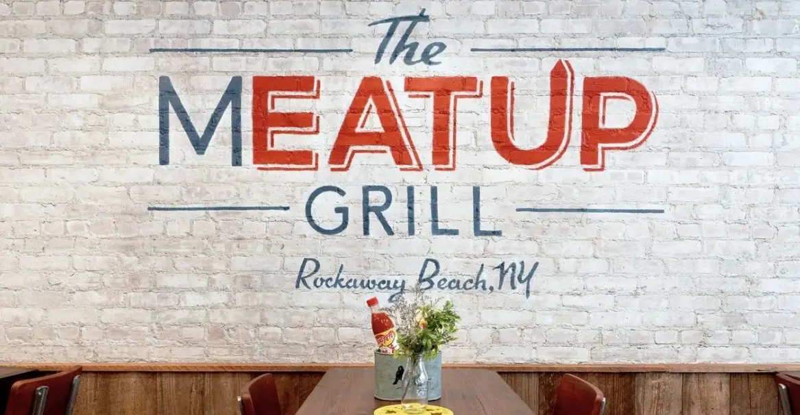Interior view of The Meat Up Grill in Rockaway Beach, NY, featuring a rustic brick wall with the restaurant's logo, creating a welcoming and cozy dining atmosphere.