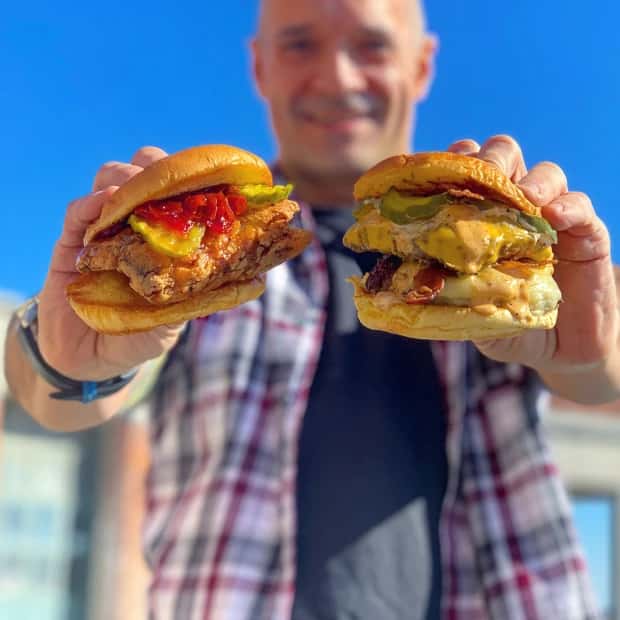 A customer at The Meat Up Grill in Rockaway Beach proudly displays two of the eatery's signature burgers, featuring the O.G. and FONZ smashed burgers, known for their elevated take on American comfort food classics.