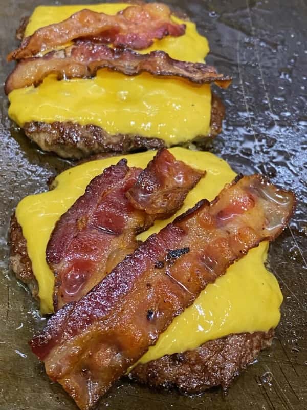 Close-up of sizzling bacon-topped cheeseburgers on the grill at The Meat Up Grill, showcasing the juicy, flavorful patties and melted cheese that define their signature American comfort food.